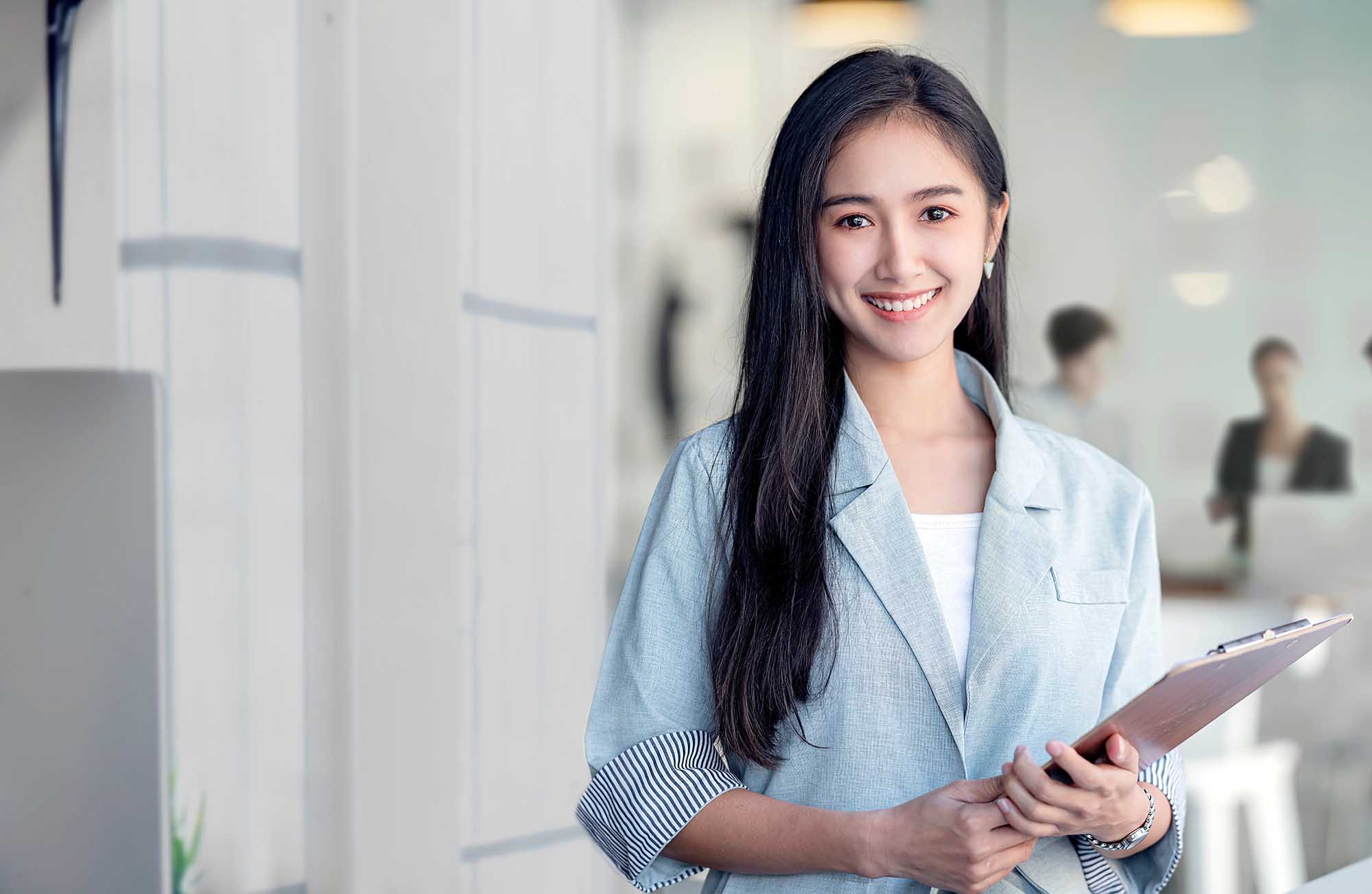 Portrait of young asian woman in casual wear holdng clipboard, smiling and looking at camera while standing in modern office room.; Shutterstock ID 1898201461; purchase_order: -; job: -; client: -; other: -